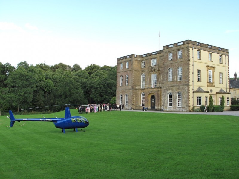 Blue helicopter G-LMBO is parked on the lawn of a large country house, group of guests in background