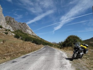 A motorcycle is parked to the side of a small country road which snakes off into hills under a crazy blue sky