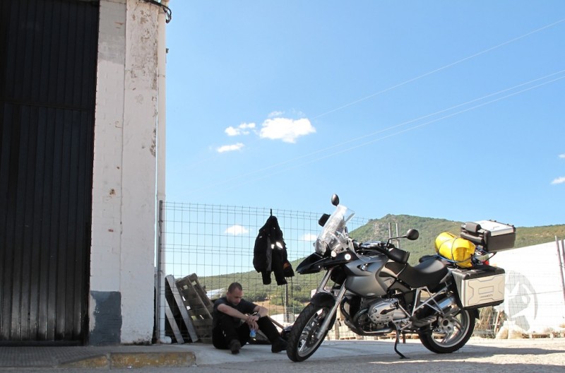 Biker sits on the floor against a fence in a petrol station, looks very hot