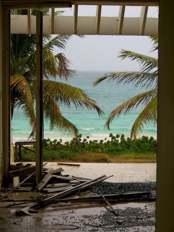 Interior view looking from ground floor apartment looking out onto beach and palm trees through broken window, parts of wooden frame occupy the floor.