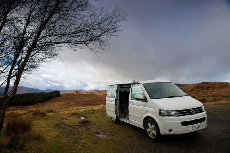 Side-on view of a white VW California camper van with side door open, parked in a Scottish valley