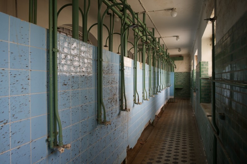 A row of bare headless showers mounted onto a cold tiled wall, splattered with unspeakable brown liquid