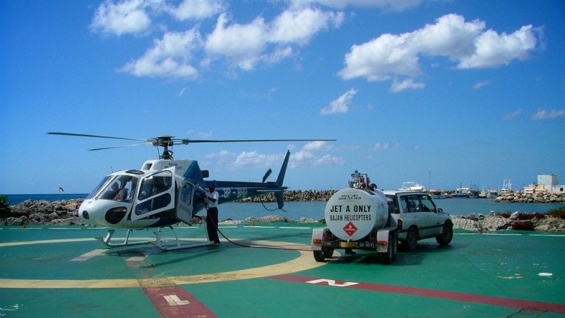 A white and blue helicopter is being refuelled on a sunny helipad under a blue sky